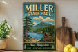 A vintage-style metal sign for Miller State Park, featuring a scenic landscape with a lake, mountains, and pine trees.