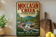 a vintage-style sign for Moccasin Creek State Park in Georgia, with a scenic illustration of a waterfall and forest. The sign encourages visitors to explore, relax, and discover the park.
