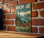 A vintage-style metal sign for Moncove Lake State Park, featuring a scenic image of a lake surrounded by mountains and a red canoe, mounted on a brick wall.