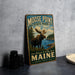 A vintage-style metal sign for Moose Point State Park, featuring a moose and a scenic landscape, is displayed on a gray countertop next to some black camera lenses.