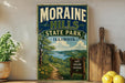 A vintage-style metal sign for Moraine Hills State Park in Illinois, featuring a scenic landscape with a path leading to a lake.