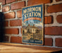 A vintage-style metal sign for Mormon Station, a historic park in Nevada, featuring an illustration of a wagon and a sign that reads "A Reminder of a Pioneer Heritage".