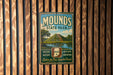 A vintage metal sign for Mounds State Park, featuring a scenic landscape with a mountain and a river, hangs on a wooden wall.