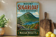 A vintage-style metal sign for Mount Sugarloaf State Reserve in Massachusetts, featuring a scenic mountain landscape with a lake and pine trees.