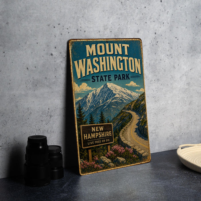 A vintage metal sign for Mount Washington State Park, featuring a mountain landscape and a winding road, is displayed on a gray countertop next to some black camera lenses.