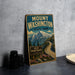 A vintage metal sign for Mount Washington State Park, featuring a mountain landscape and a winding road, is displayed on a gray countertop next to some black camera lenses.
