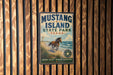 A vintage-style metal sign for Mustang Island State Park in Texas, featuring an illustration of a horse on a beach with a cloudy sky in the background.