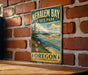 A vintage-style metal sign for Nehalem Bay State Park in Oregon, featuring a scenic landscape with a river, mountains, and pine trees.