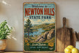 a vintage-style sign for Newton Hills State Park, with a scenic landscape of mountains, trees, and a river. The sign is displayed on a shelf next to a wooden cutting board and some potted plants.