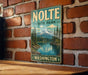 A vintage-style metal sign for NOLTE State Park in Washington, featuring a scenic mountain landscape with a lake and pine trees.