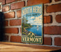 A vintage-style metal sign for Vermont State Park, featuring a scenic landscape with a sailboat on the water and a mountain in the background.