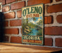 A vintage-style sign for O'Leno State Park in Florida, featuring a scenic landscape with a river and palm trees.