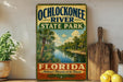 A vintage-style sign for Ochlockonee River State Park in Florida, featuring a tropical landscape with palm trees and a river.
