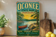a vintage-style sign for Oconee State Park, South Carolina, with a scenic illustration of a lake, trees, and a canoe. The sign also includes the words "Nature, Adventure, Memories, Explore, Relax, Discover" in a green and yellow color scheme.