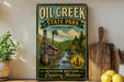 a vintage-style sign for Oil Creek State Park in Pennsylvania, with a scenic illustration of a river, a steam engine, and a wooden building. The sign is displayed on a shelf next to a wooden cutting board and a potted plant.