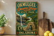 A vintage-style sign for Okmulgee-Drip Springs State Park in Oklahoma, featuring a waterfall and a stone structure.