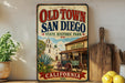 A vintage-style metal sign for Old Town San Diego, California, featuring an illustration of a historic building and a wagon, with a wooden paddle and a potted plant in the foreground.