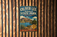 A vintage-style metal sign for Oliver Lee Memorial State Park in New Mexico, featuring a scenic landscape with mountains, a lake, and desert vegetation.