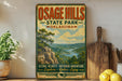 a vintage-style poster for Osage Hills State Park in Oklahoma, with a scenic mountain landscape and a river. The poster is displayed on a wall, accompanied by a wooden cutting board and a potted plant.