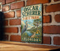 A vintage-style metal sign for Oscar Scherer State Park in Florida, featuring a blue heron and palm trees, is displayed on a brick wall.
