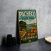 A vintage metal sign for Pacheco State Park, featuring a scenic landscape with a path, trees, and flowers, is displayed on a gray countertop next to a stack of black camera lenses.