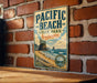 A vintage-style metal sign for Pacific Beach State Park in Washington, featuring a scenic coastal landscape with a beach, mountains, and a body of water.