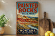 A vintage-style metal sign for Painted Rocks State Park in Montana, featuring a scenic landscape with mountains, a river, and a wooden paddle.