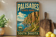 A vintage-style metal sign for Palisades State Park in South Dakota, featuring a scenic mountain landscape with a lake and trail.
