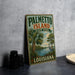 A vintage-style metal sign for Palmetto Island State Park in Louisiana, featuring a palm tree and a bird, is displayed on a gray countertop next to a stack of black camera lenses.