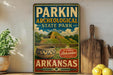A vintage-style sign for Parkin Archaeological State Park in Arkansas, featuring a pottery bowl and a wooden paddle, is displayed on a shelf next to a potted plant and some lemons.