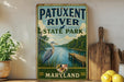 A vintage-style metal sign for Patuxent River State Park in Maryland, featuring an illustration of a heron and a wooden dock on a river.