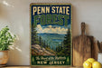 A vintage-style sign for Penn State Forest, featuring a scenic mountain landscape with pine trees and a lake, is displayed on a wooden shelf next to a potted plant and a cutting board.