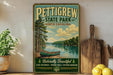 A vintage-style sign for Pettigrew State Park, featuring a scenic image of a lake with a canoe, surrounded by pine trees and mountains.
