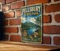 A vintage metal sign for Pillsbury State Park in New Hampshire, featuring a scenic mountain landscape and inviting visitors to swim, hike, explore, relax, and enjoy nature.