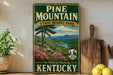 A vintage-style sign for Pine Mountain State Resort Park in Kentucky, featuring a scenic mountain landscape with pine trees, a lake, and mountains in the background.