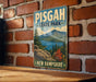 A vintage-style metal sign for Pisgah State Park in New Hampshire, featuring a scenic landscape with mountains, pine trees, and a lake.