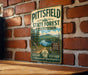 A vintage metal sign for the Pittsfield State Forest in Massachusetts, featuring a scenic landscape with pine trees, a lake, and mountains.