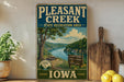 A vintage-style sign for Pleasant Creek State Recreation Area in Iowa, featuring a scenic image of a river and mountains.