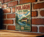 A vintage-style metal sign for Pokagon State Park, Indiana, featuring a scenic illustration of a bridge and a river, mounted on a brick wall.