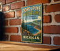 A vintage-style metal sign for Porcupine Mountains Wilderness State Park in Michigan, featuring a scenic mountain landscape with pine trees and a lake.