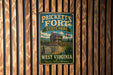 A vintage metal sign for Prickett's Fort State Park in West Virginia, featuring a cabin and a flag, mounted on a wooden wall.