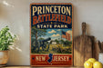 A vintage metal sign for Princeton Battlefield State Park in New Jersey, featuring an illustration of a cannon and a field with a building and an American flag.