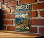 A vintage metal sign for the Prospect State Scenic Viewpoint in Oregon, featuring a mountain landscape and a stone structure.