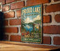 A vintage metal sign for "Proud Lake Recreation Area" is displayed on a brick wall, featuring a scenic image of a lake and a canoe.