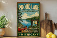 A vintage-style metal sign for "Proud Lake Recreation Area" in Michigan, featuring a scenic image of a lake, canoe, and surrounding forest.