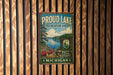 A vintage metal sign for "Proud Lake Recreation Area" in Michigan, featuring a scenic image of a lake, canoe, and surrounding forest.