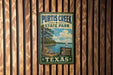 A vintage metal sign for Purtis Creek State Park in Texas, featuring a scenic landscape with a lake, trees, and a sailboat.