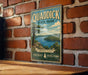 A vintage-style sign for Quaddick State Park, featuring a scenic landscape with a lake and mountains, mounted on a brick wall.