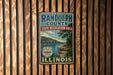A vintage metal sign for Randolph County, Illinois, featuring a scenic landscape with a lake and mountains.