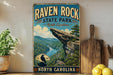 A vintage-style sign for Raven Rock State Park in North Carolina, featuring a scenic mountain landscape with a river and a bird perched on a rock.